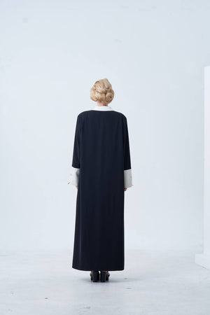 Person wearing a long black dress with white cuffs and hem against a white background
