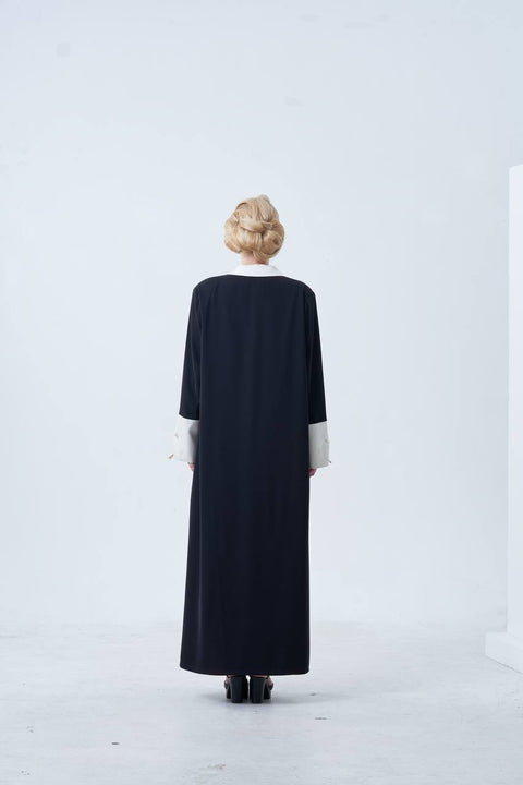 Person wearing a long black dress with white cuffs and hem against a white background